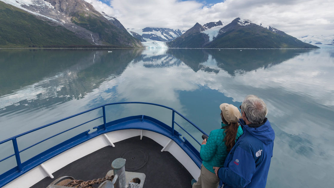 Couple stands on bow of blue-&-white Alaska small ship Discovery, in calm waters on a partly sunny day.