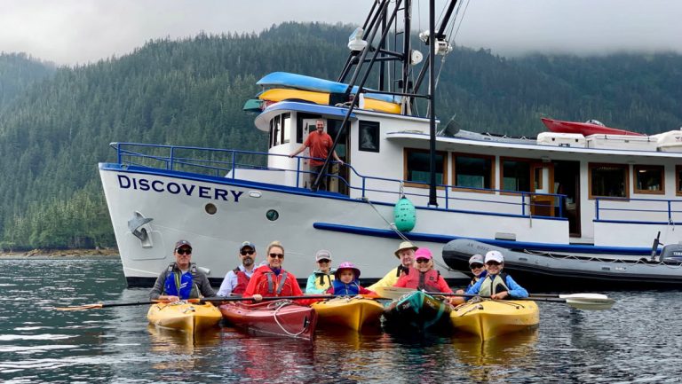 Family of kayakers sits in yellow boats in the water with Alaska small ship Discovery behind, on a cloudy day.