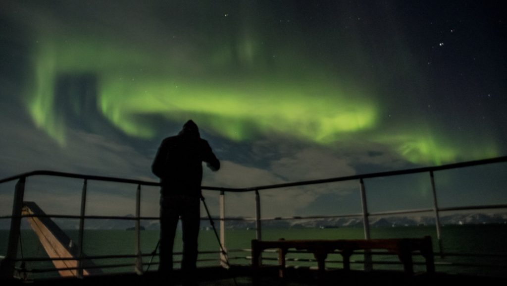 Person stands in deck of a ship at night, watching the green aurora borealis on the Scoresby Sund Aurora Borealis voyage.