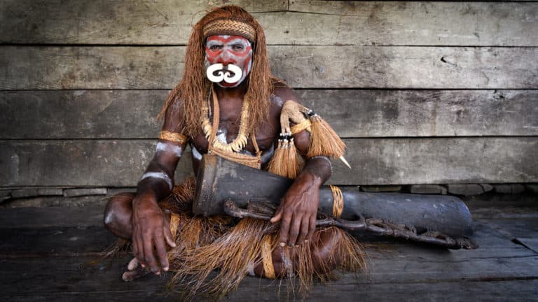 West Papuan tribesman sits with a wooden drum in front of a weathered wood-plank wall during the Spice Islands & Raja Ampat small ship cruise in Indonesia.