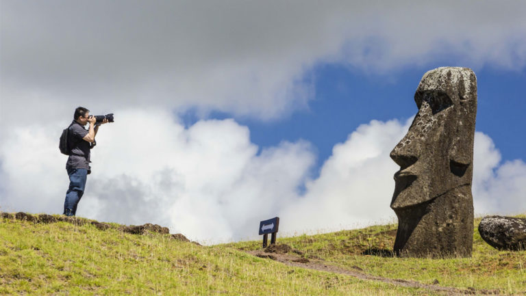 solo traveler takes a photograph of a rapa nui statue in easter island chile while standing on a grassy knoll on a partly sunny day