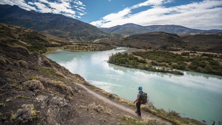 Man stands on smooth dirt trail overlooking a milky turquoise glacial lake on a sunny day during the Torres Del Paine Trek.