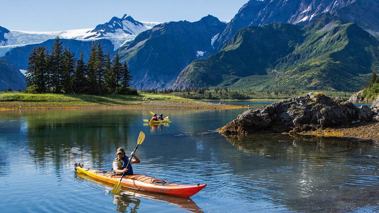 Kayakers in yellow & orange boats paddle glassy water beside tall green mountains on the Ultimate Alaska Adventure.