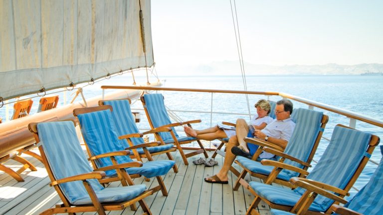 Man & woman sit on teak deck chairs lined with bright blue padding, reading under full sail of the Sea Cloud ship in the Mediterranean.