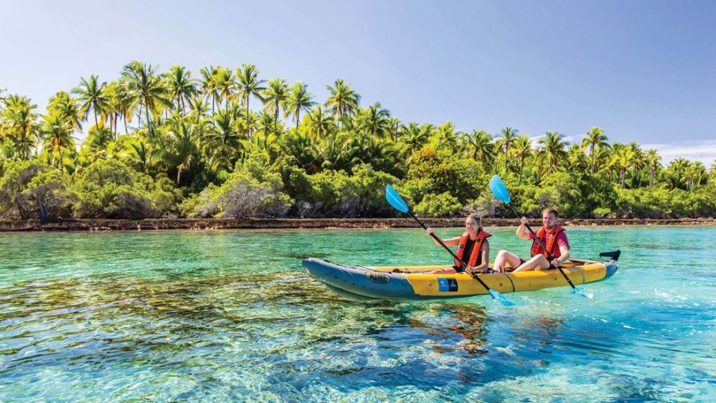 A girl and boy paddle a yellow kayak in crystal clear teal water front of a palm tree filled sandy island in Polynesia.