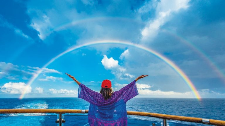 Woman stands at bow of ship & raises both arms toward a double rainbow over the sea on a sunny day near Easter Island.