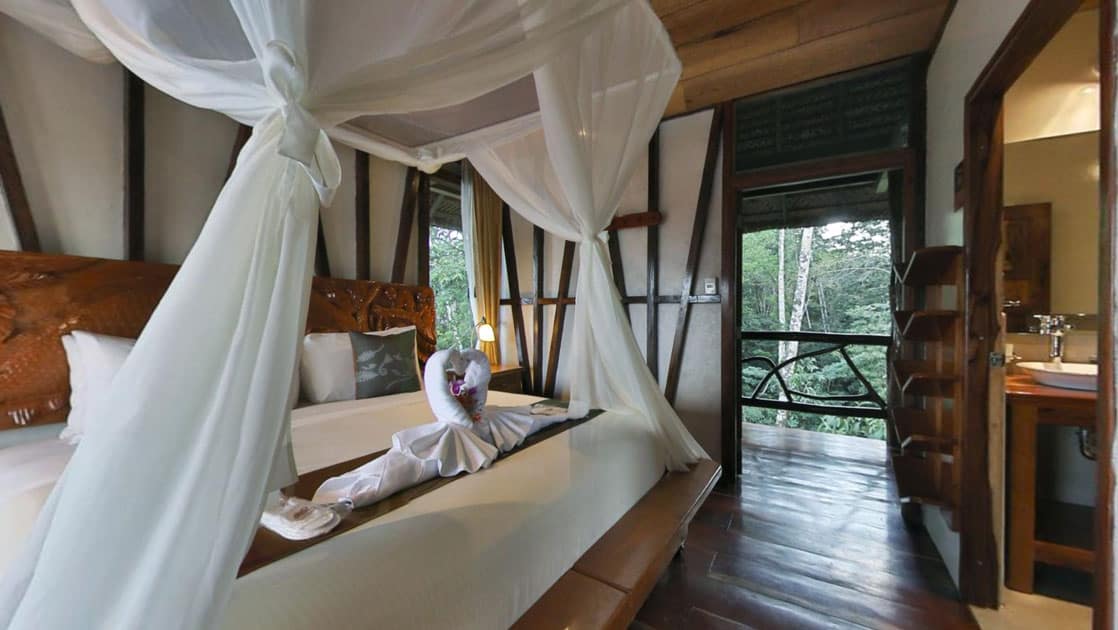 Large bed with carved wooden headboard, white sheets & white canopy looks onto bathroom and open door to the rainforest.