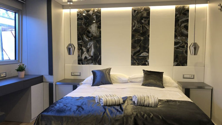 Double bed with white & black bedding & dark marble headboard beside bright window in a cabin on Aurelia deluxe Mediterranean yacht.