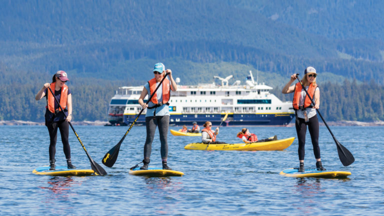 Three women stand-up paddleboarding with a National Geographic expedition ship in the background, on a sunny day.