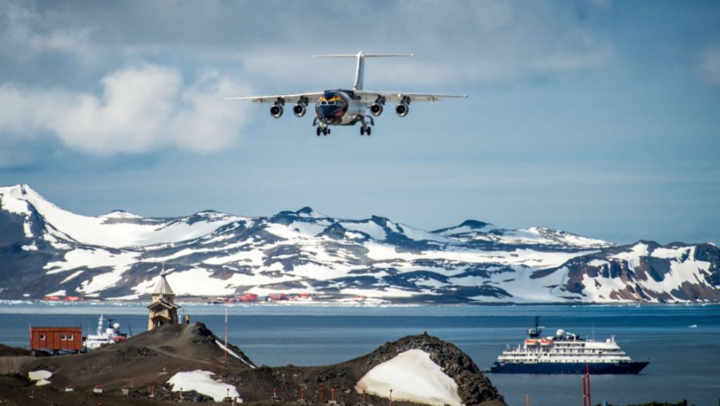 For an express fly cruise itinerary an Antarctica expedition ships floats in the ocean while a charter plane flies through the blue sky headed to the landing strip below.