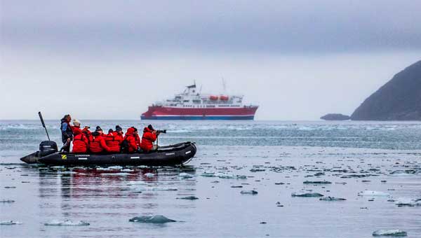 Arctic cruise travelers in red jackets take photographs of iceberg bits in the water on a Zodiac cruise to the Arctic Circle, with a small polar ship in the background.