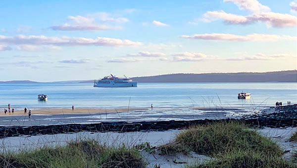 Australia small ship cruise passengers walk on the shoreline with the ship at anchor in the bay and two skiffs in the water