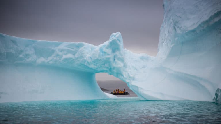 A Zodiac pauses in the distance, framed by a hole in an iceberg, on an overcast day, during the Greenland Adventure cruise by land, sea and air.