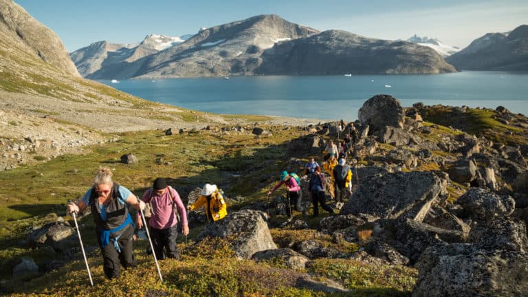 A line of hikers walk over rocky, grassy knolls, on a sunny day, during the Greenland Adventure cruise by land, sea and air.