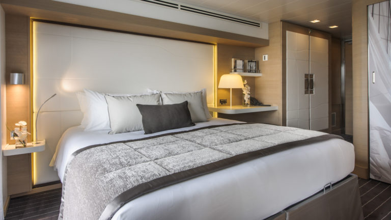 Superior Stateroom with king bed aboard Le Soleal luxury expedition ship, caramel colored walls with white bedding and headboard and featuring grey accent pillows