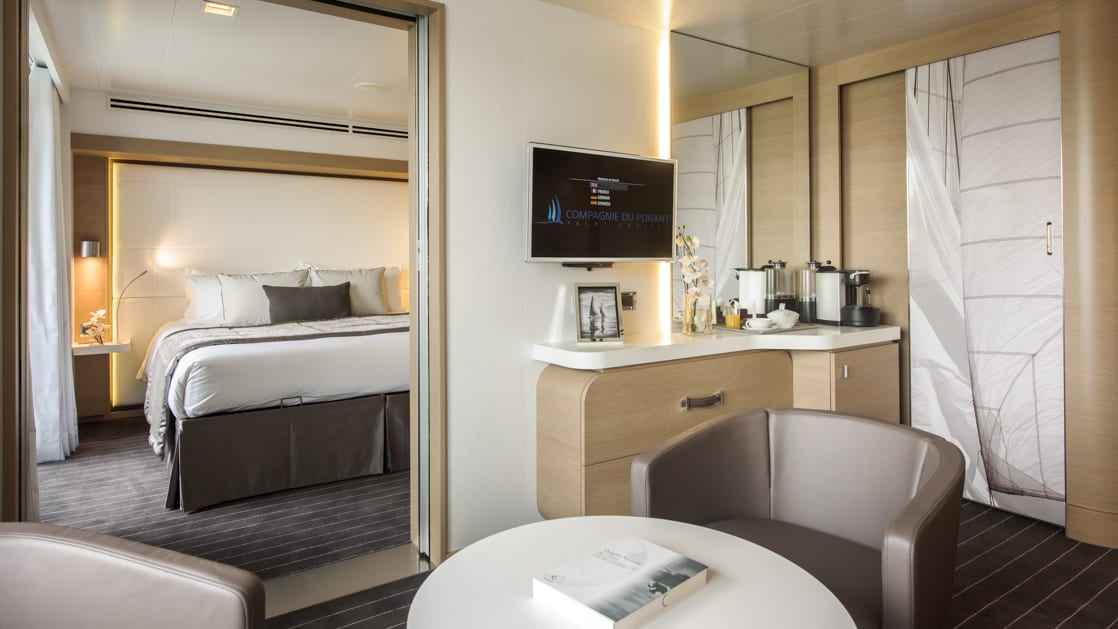 Prestige Suite with king bed aboard Le Soleal, a grey and white seating area with a flat screen tv on the wall.