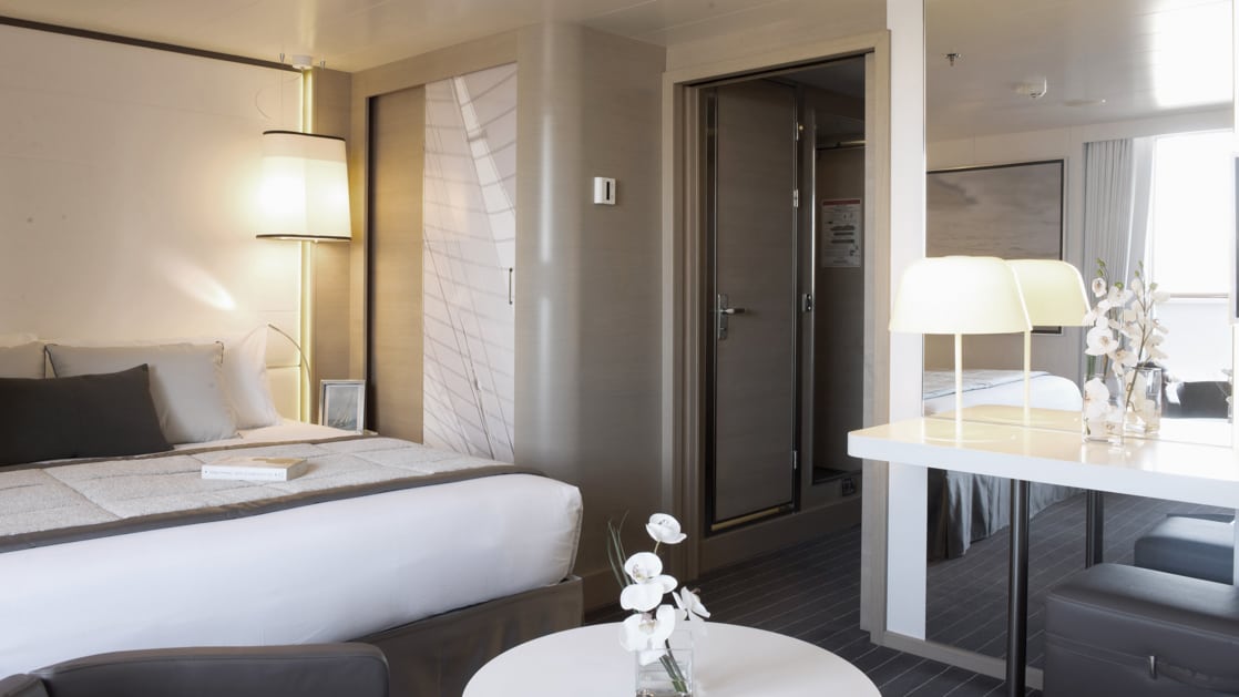 Deluxe Suite with king bed aboard Le Soleal white and grey bedding and decor with a coffee table and closet.