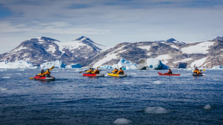5 tandem paddlers travel on icy blue waters with snowy mountains in the background during The Northwest Passage Canadian High Arctic voyage.