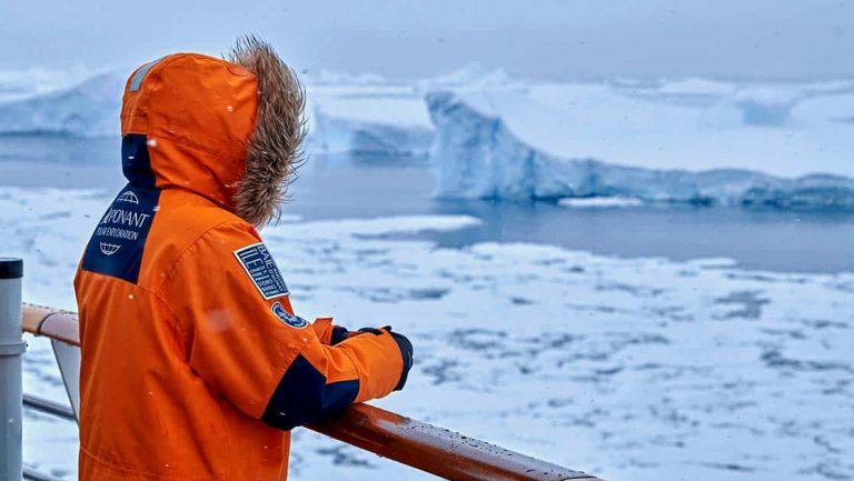 An Antarctica cruise guest leans on the ship railing wearing an orange parka with fur hood looking out over an icy landscape.