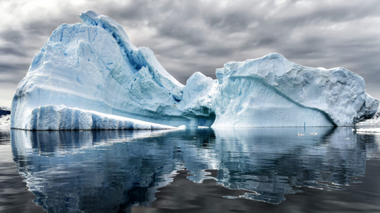 A pair of large icebergs reflect icy blue into glassy waters during the Great Austral Loop luxury Antarctica voyage.