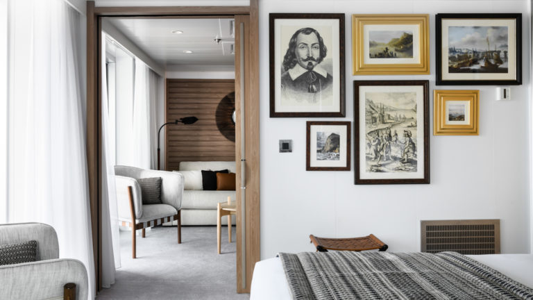 Owner's Suite aboard Le Champlain luxury expedition ship, showing king bed, separate living room, bright white decor & photos & drawings of explorers on the walls.