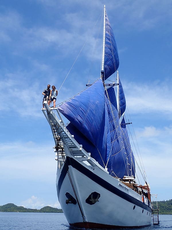two cruise guests stand high above the water on the long wooden bow of the ombak putih, they lean against a rope from its sails