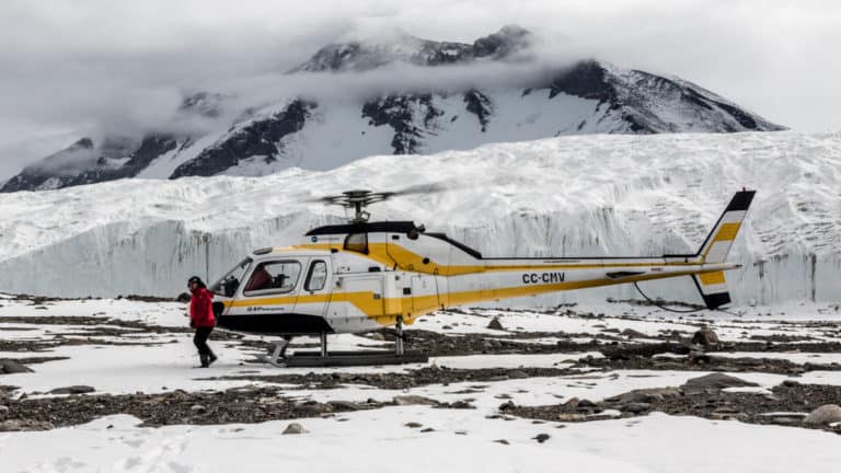 A helicopter sits on land while a guest walks around the snowfield at Taylor Valley, Dry Valleys, McMurdo Sound, during the Spectacular Ross Sea: West Antarctica Cruise.