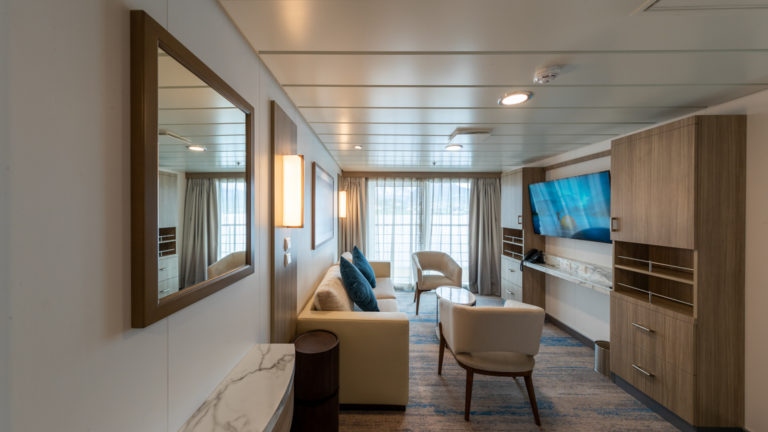 Lounge area of Captain's Suite aboard Greg Mortimer polar ship, with framed mirror on the all, wardrobe, beige couch & 2 chairs, flatscreen TV & sliding glass doors out onto private balcony.