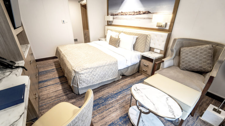 Junior Suite aboard Greg Mortimer polar ship, with beige, quilted king bed, gray chair, marble side table, desk, drawers & framed Antarctica photo with reading lights above headboard.