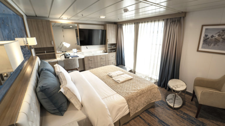 Superior Balcony Stateroom aboard Greg Mortimer polar ship, with beige quilted king bed, marble side table, tan chair, flatscreen TV & sliding glass doors onto private balcony.