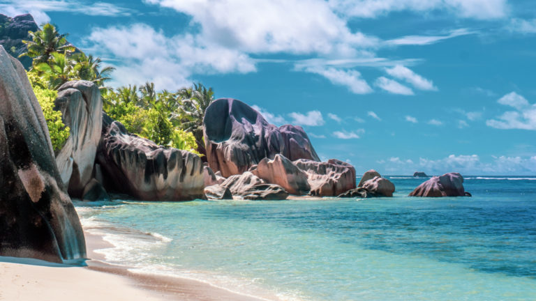 Private beach lined with red boulders & palm trees on a sunny day during the Pearls of the Caribbean luxury cruise.