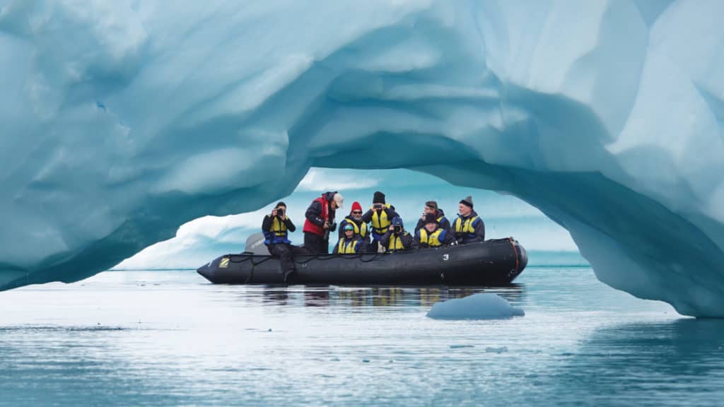 A group of polar travelers in a Zodiac cruises beside an archway in a large blue iceberg during the Spirit of Antarctica expedition.