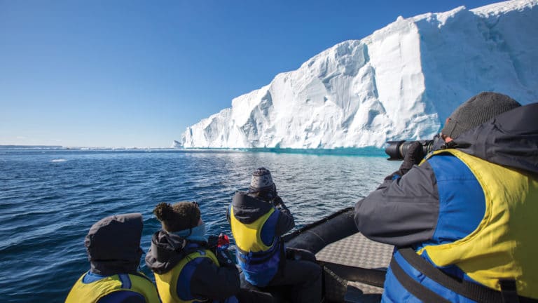 A group of polar travelers in yellow life jackets sit in a Zodiac and photograph a large tabular iceberg on a sunny day during the Active & Wild Antarctica Air Cruise.