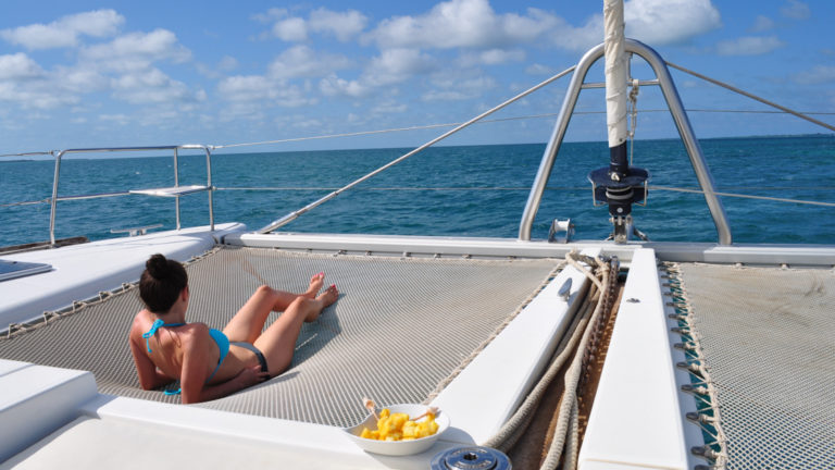 Woman in a bikini lounges on the trampoline netting at the front of her private Belize catamaran charter on a sunny day.