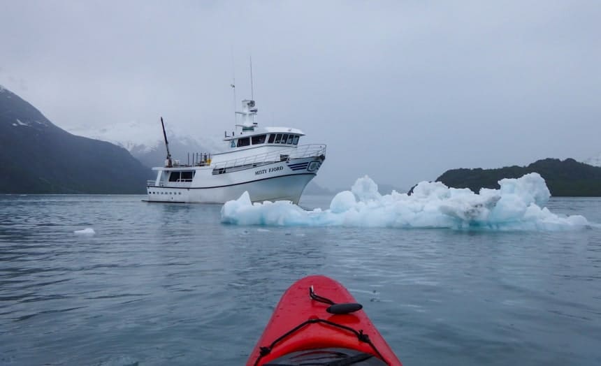 The tip of a red kayak floats in the foreground, infront of it floats an iceberg and the Misty Fjord ship on the islands whales glaciers cruise.
