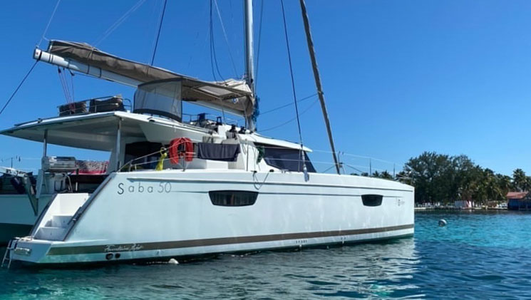 White catamaran named Nowhere sits in turquoise water with a small island in the background on a sunny day in Belize.