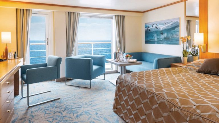 Owner's Suite aboard Seaventure Antarctica small ship, with queen bed covered in brown bedspread, sitting area with couch & chairs, & large balcony.