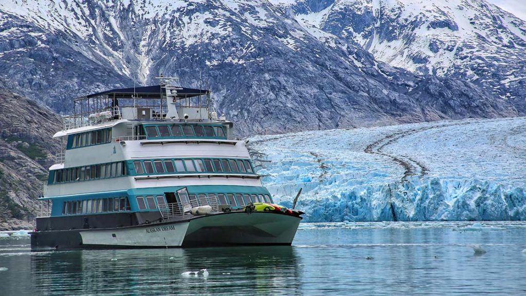 The white and teal Alaskan Dream ship cruises in front of a large icy teal glacier flanked by a rocky mountain range.