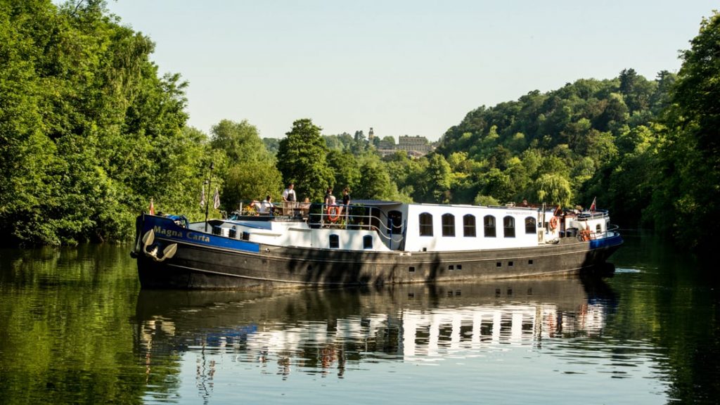 Black, white & blue-painted Magna Carta barge cruises the River Thames in calm waters with tree-lined riverbanks.