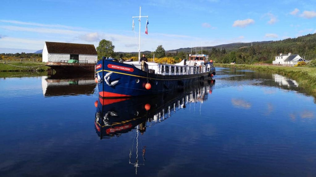 Scottish Highlander hotel barge sitting still in calm water with red-&-blue exterior & small white wooden building behind, on a sunny day in Scotland.