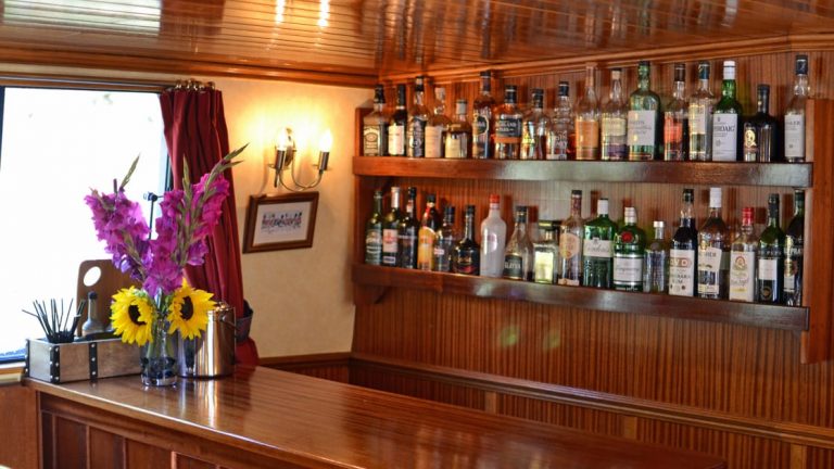 Warm wooden bar with 2 shelves of whisky behind & view window with vase of flowers beside it, aboard Scottish Highlander hotel barge.