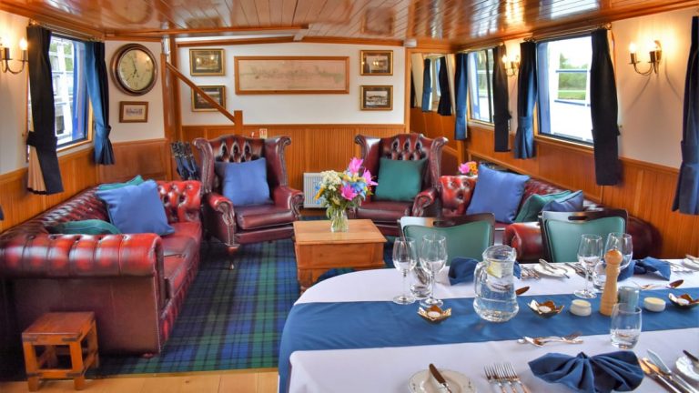 Saloon indoor common area with dining table set for service & living room behind with leather armchairs & sofas aboard Scottish Highlander Caledonia Canal small ship.