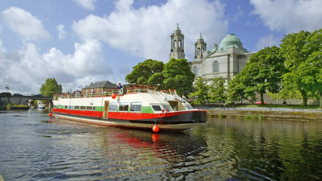 Shannon Princess barge cruising Ireland's River Shannon, past a castle & bright green trees on a sunny day.