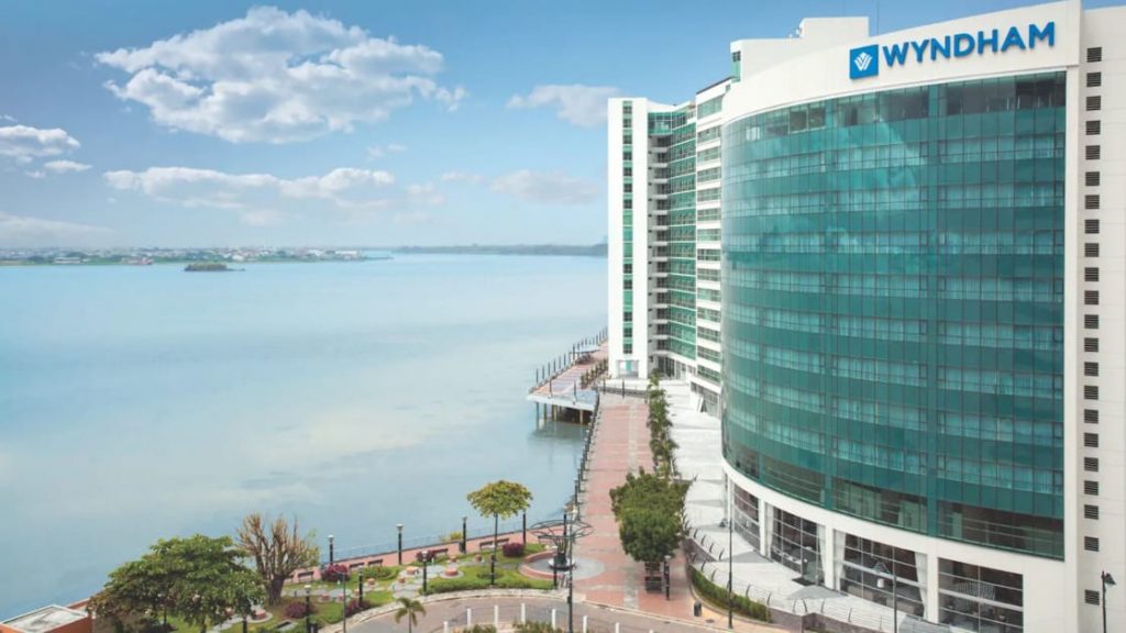 Exterior of Wyndham Guayaquil Hotel, multiple-story glass-encased building overlooking a large scenic river and a riverside walkway on a sunny day.