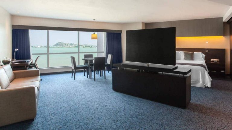 Junior Suite at Wyndham Guayaquil Hotel, with panoramic, floor-length windows, dresser & flat-screen TV separating bedroom from living area, table & chairs & double bed.