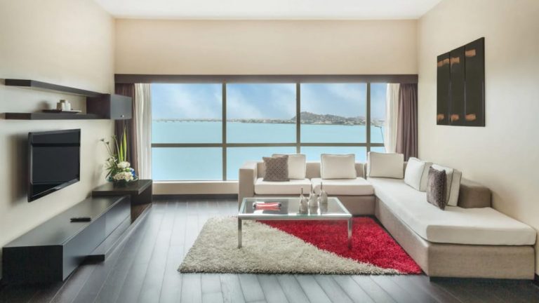 Presidential Suite living room at Wyndham Guayaquil Hotel with wraparound white couch, floor-length windows, flat-screen TV, coffee table, red-&-white rug & dark wooden flooring.