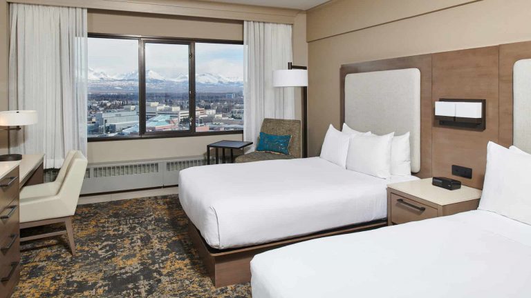 A double bed room inside the Hilton Downtown Anchorage. Two separate beds, white comforters, chair with ottoman and floor lamp city view windows.