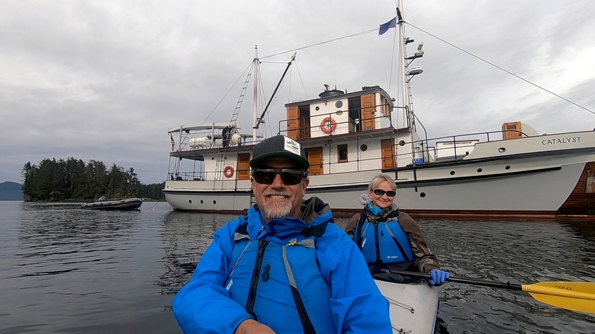 Two cruise guests a male and female, sit inside a double kayak together and paddle around the Alaska small ship Catalyst.