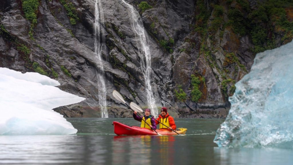 Two travelers paddle a double kayak between icebergs and in front of a waterfall on an Alaska small ship cruise.