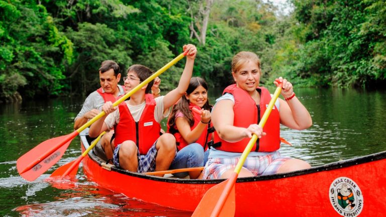 Family smiling while paddling a red canoe on a river in the jungle in Belize on a tour.